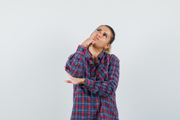 Giovane donna in piedi nella posa di pensiero mentre mette il dito indice sul mento in camicia a quadri e guardando pensieroso, vista frontale.