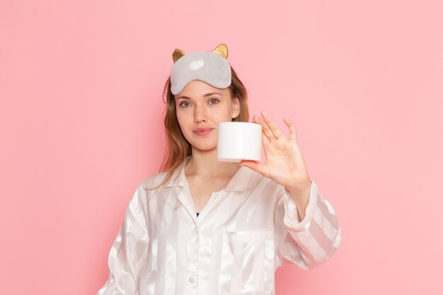 giovane donna in pigiama e maschera per dormire tenendo la crema con il sorriso sul rosa