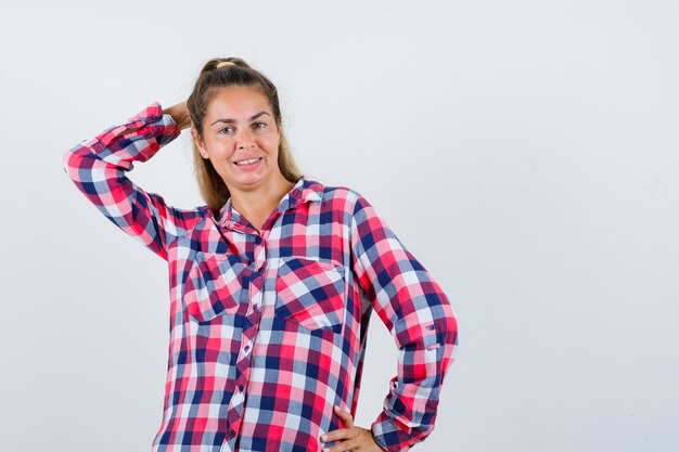 Giovane donna in posa con la mano dietro la testa in camicia a quadri e guardando allegro, vista frontale.