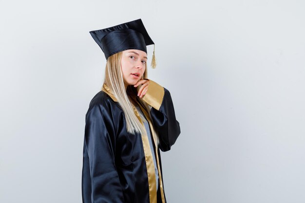 Giovane donna in posa con la mano sul mento in uniforme da laureato e dall'aspetto grazioso