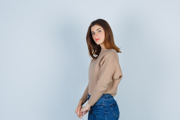 Giovane donna in posa mentre guarda davanti in maglione beige, jeans e sembra sicura, vista frontale.