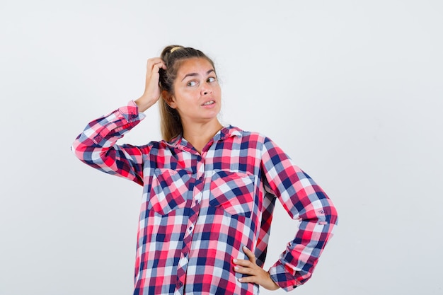 Giovane donna in posa mentre si gratta la testa in camicia a quadri e sembra ragionevole. vista frontale.