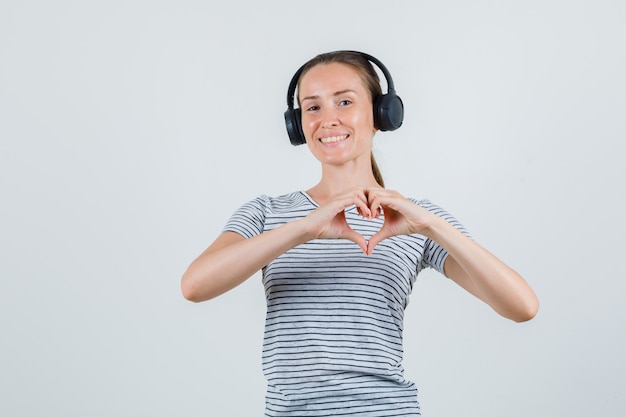 Giovane donna in t-shirt, cuffie che mostrano il gesto del cuore e che sembra allegro, vista frontale.