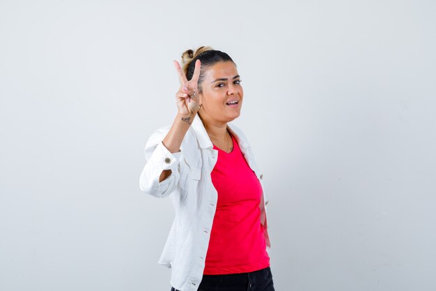 Giovane donna in t-shirt, giacca bianca che mostra il segno della vittoria e sembra gioiosa, vista frontale.