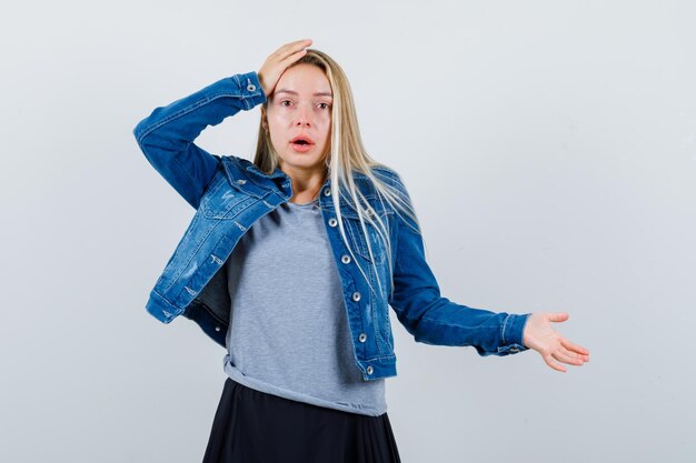 Giovane donna in t-shirt, giacca di jeans, gonna che tiene la mano sulla testa e sembra impotente