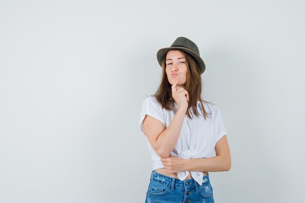 Giovane donna in t-shirt, jeans, cappello in piedi nella posa di pensiero e che sembra sensibile, vista frontale.