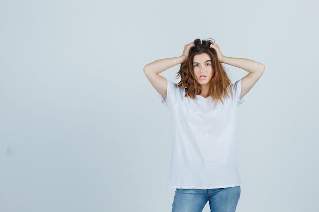 Giovane donna in t-shirt, jeans che tengono le mani sulla testa e guardando stupito, vista frontale.