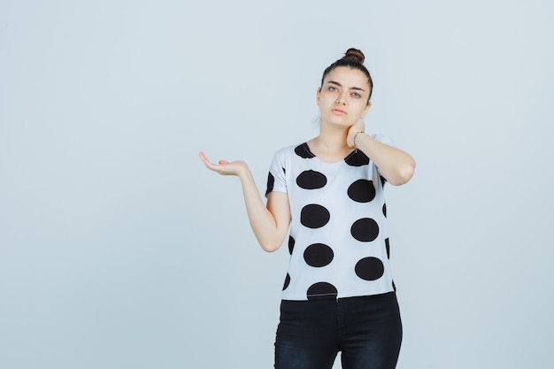 Giovane donna in t-shirt, jeans tenendo la mano sul collo fingendo di tenere qualcosa e guardando pensieroso, vista frontale.