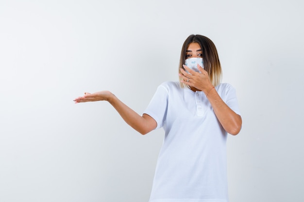 Giovane donna in t-shirt, maschera che dà il benvenuto a qualcosa tenendo la mano sulla bocca e guardando fiducioso, vista frontale.