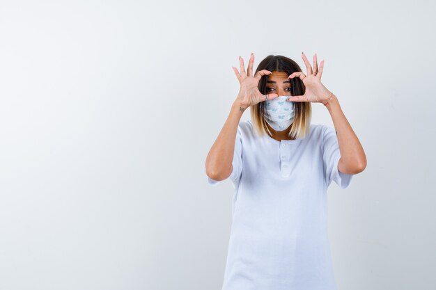 Giovane donna in t-shirt, maschera che mostra il gesto degli occhiali e che sembra carina, vista frontale.