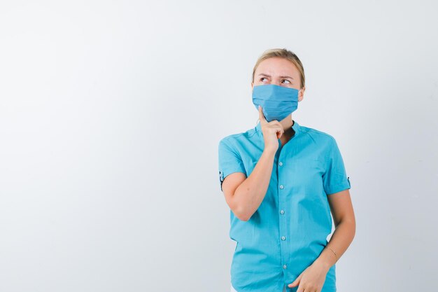 Giovane donna in t-shirt, maschera in piedi in posa pensante e dall'aspetto pensieroso