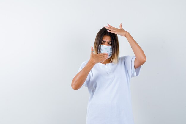 Giovane donna in t-shirt, maschera tenendo la mano sulla bocca mantenendo la mano sulla testa e guardando esitante, vista frontale.