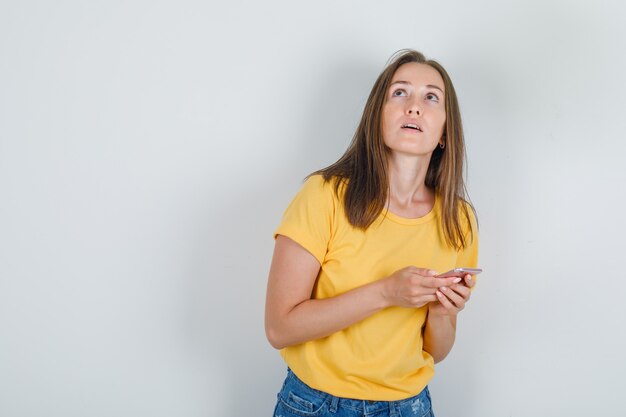 Giovane donna in t-shirt, pantaloncini alzando lo sguardo mentre si tiene lo smartphone e guardando pensieroso