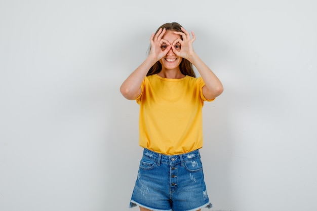 Giovane donna in t-shirt, pantaloncini che mostrano il gesto degli occhiali e che sembra allegra