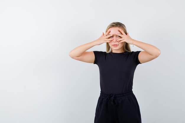 Giovane donna in t-shirt, pantaloni che guarda attraverso le dita e che sembra spaventata, vista frontale.