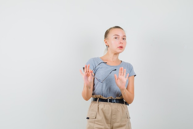 Giovane donna in t-shirt, pantaloni che mostra il gesto di rifiuto mentre si tengono gli occhiali, vista frontale.