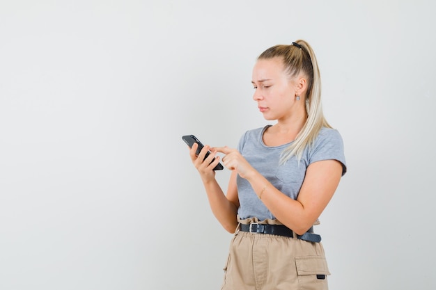 Giovane donna in t-shirt, pantaloni utilizzando il telefono cellulare e guardando occupato, vista frontale.