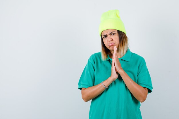 Giovane donna in t-shirt polo, berretto con le mani in gesto di preghiera e sguardo sconvolto, vista frontale.