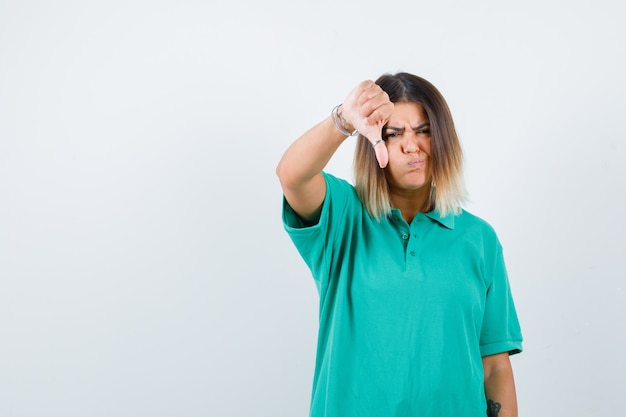 Giovane donna in t-shirt polo che mostra il pollice verso il basso con le guance gonfie e sembra insoddisfatta, vista frontale.