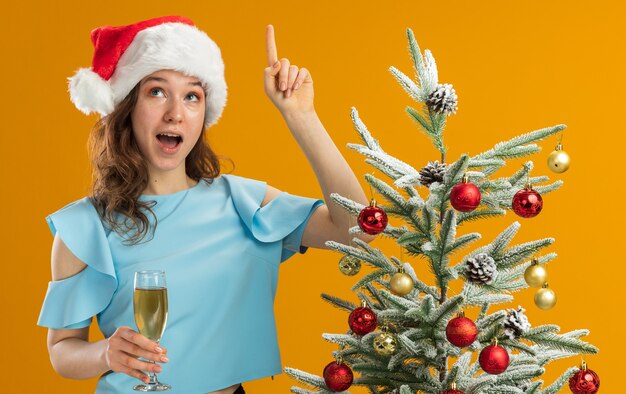 Giovane donna in top blu e cappello da Babbo Natale con in mano un bicchiere di champagne alzando lo sguardo sorpreso mostrando il dito indice che ha una nuova idea in piedi accanto a un albero di Natale sul muro arancione