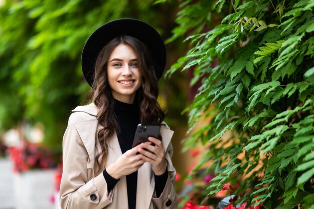 Giovane donna in un cappello cammina in città e utilizza uno smartphone. Hipster in una passeggiata utilizza il telefono e scatta foto per i social network