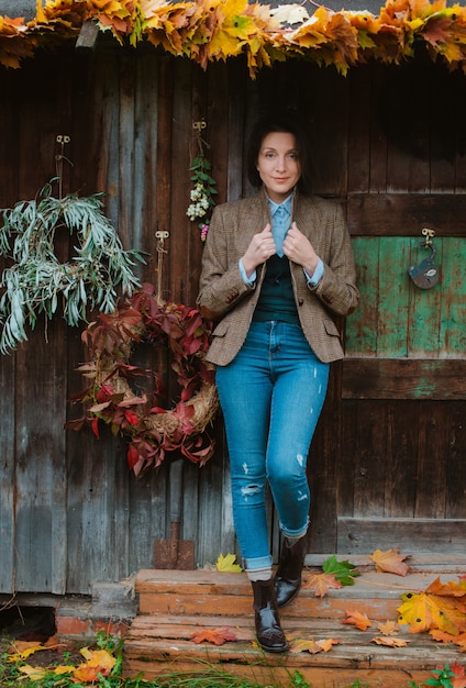 Giovane donna in una giacca calda marrone e jeans pone su una casa rustica