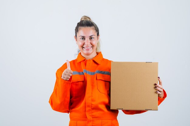 Giovane donna in uniforme da lavoro che mostra il pollice in su mentre si tiene la scatola e guardando allegro, vista frontale.