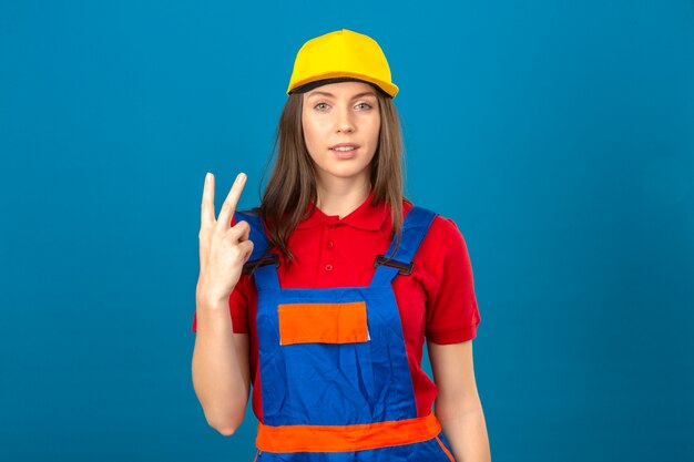 Giovane donna in uniforme di costruzione e casco di sicurezza giallo che esaminano macchina fotografica che mostra numero due con le dita che stanno sul fondo blu