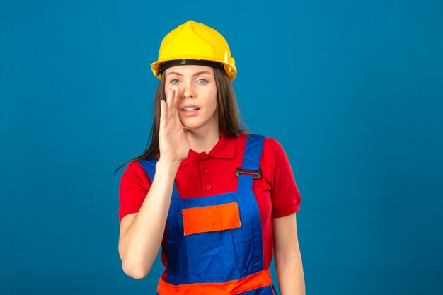 Giovane donna in uniforme di costruzione e casco di sicurezza giallo con una bocca vicina della mano che dice una condizione segreta sul fondo blu