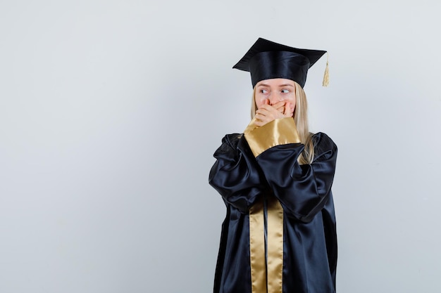 Giovane donna in uniforme laureata che tiene le mani sulla bocca e sembra spaventata