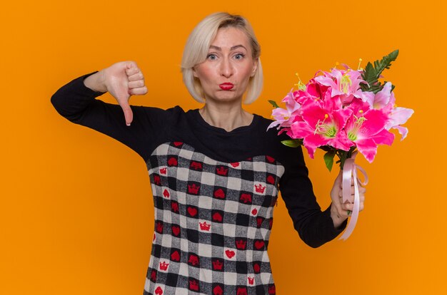 Giovane donna in vestito bello che tiene il mazzo di fiori che guarda davanti con l'espressione triste che mostra i pollici giù per celebrare la giornata internazionale della donna in piedi sopra la parete arancione