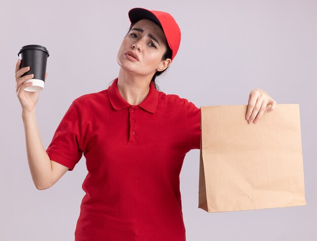Giovane donna incapace di consegna in uniforme e berretto che tiene in mano una tazza di caffè in plastica e un pacchetto di carta