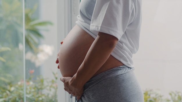 Giovane donna incinta asiatica che tiene la sua pancia che parla con suo figlio. Mamma sentirsi felici sorridenti positivi e pacifici mentre abbi cura del bambino, gravidanza vicino alla finestra nel salotto di casa.