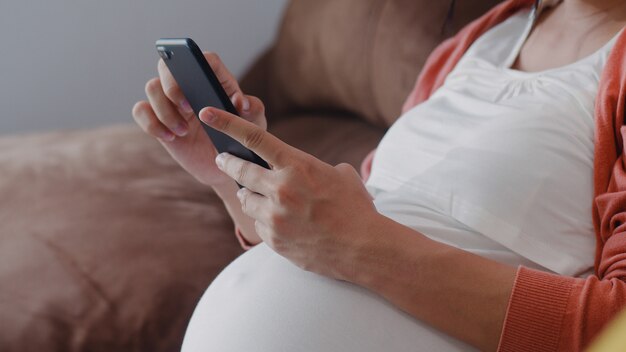 Giovane donna incinta asiatica che usando le informazioni di gravidanza di ricerca del telefono cellulare. La mamma si sente felice sorridente positivo e pacifico mentre si prende cura del suo bambino sdraiato sul divano nel salotto di casa.