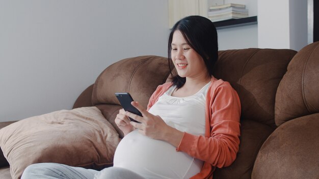 Giovane donna incinta asiatica che usando le informazioni di gravidanza di ricerca del telefono cellulare. La mamma si sente felice sorridente positivo e pacifico mentre si prende cura del suo bambino sdraiato sul divano nel salotto di casa.