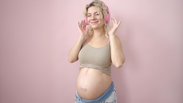 Giovane donna incinta che sorride fiduciosa ascoltando musica su sfondo rosa isolato