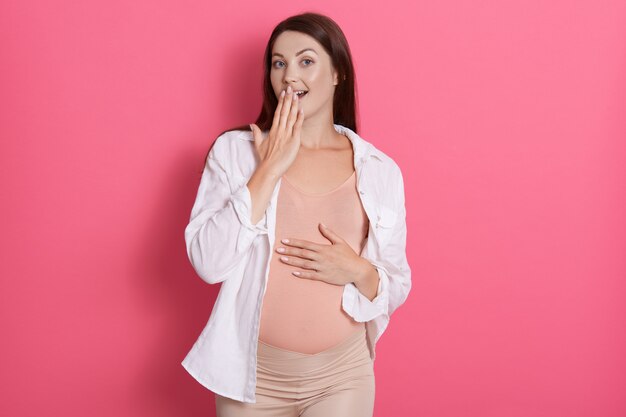 Giovane donna incinta con l'espressione di dimenticare qualcosa di isolato nello spazio rosa, tenendo la mano sulla pancia, indossando leggins e camicia casual.