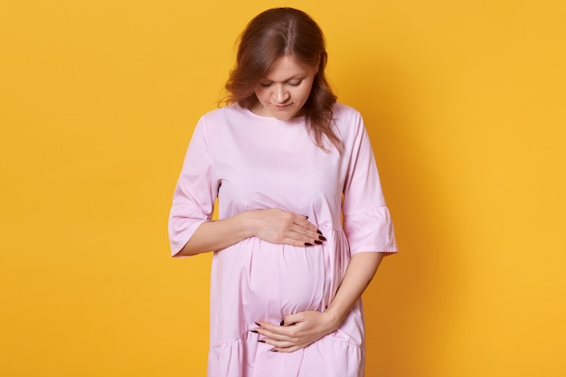 giovane donna incinta isolata su giallo, adorabile signora sta con le mani sul suo addome, guarda in basso sul ventre, immagina il suo futuro bambino. Concetto di maternità