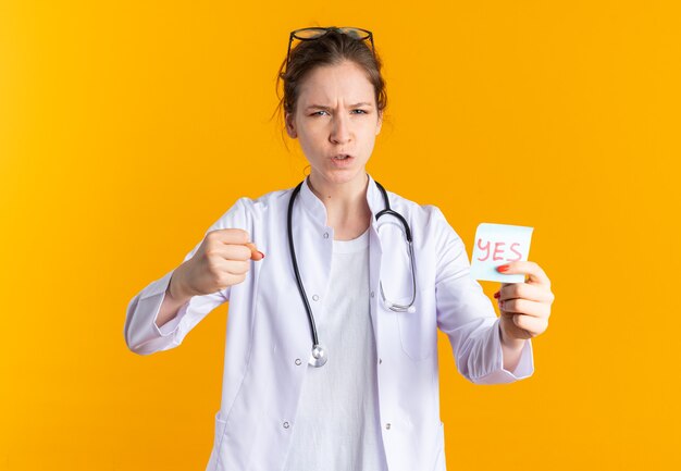 Giovane donna infastidita in uniforme da medico con stetoscopio che tiene nota di sì e tiene il pugno
