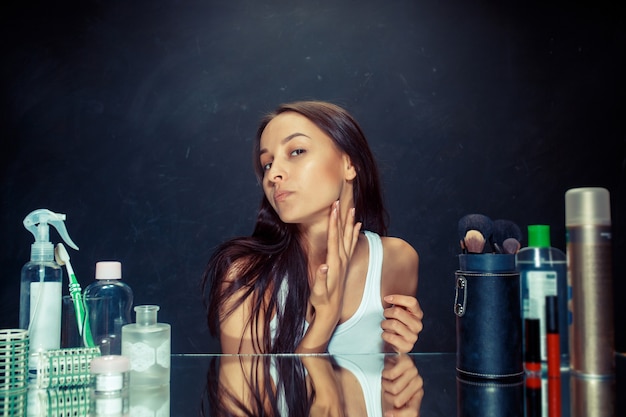 Giovane donna infelice insoddisfatta che esamina la sua auto in specchio su sfondo nero studio. concetto di pelle e acne roblem. Mattina, trucco e concetti di emozioni umane. Modello caucasico in studio