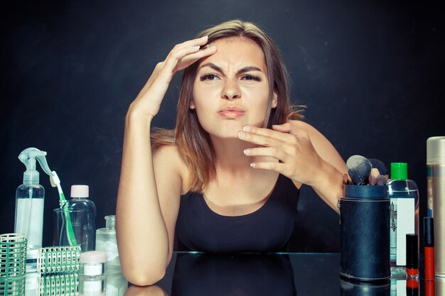 Giovane donna infelice insoddisfatta che esamina la sua auto in specchio su sfondo nero studio. concetto di pelle e acne roblem. Mattina, trucco e concetti di emozioni umane