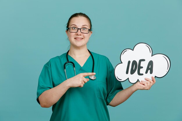 Giovane donna infermiera in uniforme medica con lo stetoscopio intorno al collo che tiene il discorso della bolla con l'idea di parola che punta con il dito indice su di essa sorridente sorpreso in piedi su sfondo blu