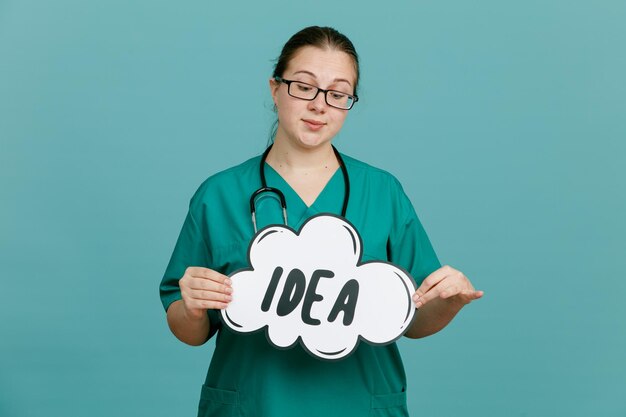Giovane donna infermiera in uniforme medica con lo stetoscopio intorno al collo che tiene il discorso della bolla con l'idea di parola che sembra sicura felice e positiva in piedi su sfondo blu