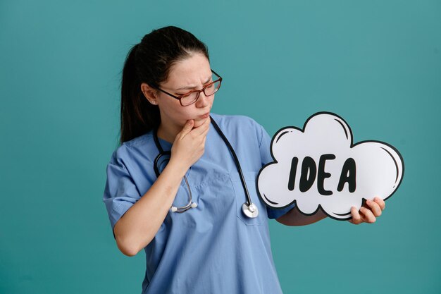 Giovane donna infermiera in uniforme medica con lo stetoscopio intorno al collo che tiene il fumetto con l'idea di parola guardandolo con espressione pensierosa che pensa in piedi su sfondo blu