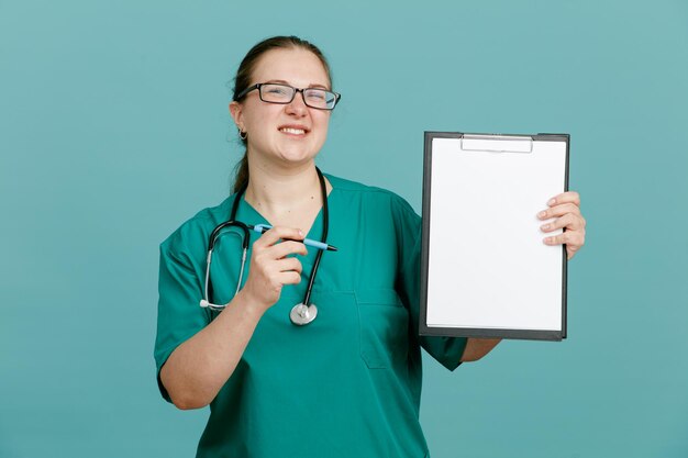 Giovane donna infermiera in uniforme medica con stetoscopio intorno al collo che tiene appunti e penna guardando la fotocamera felice e positivo sorridente fiducioso in piedi su sfondo blu