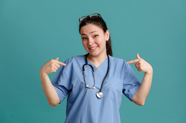Giovane donna infermiera in uniforme medica con stetoscopio intorno al collo guardando la fotocamera felice e soddisfatto di sé che punta a se stessa in piedi su sfondo blu