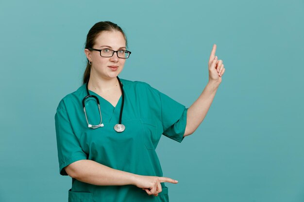 Giovane donna infermiera in uniforme medica con stetoscopio intorno al collo guardando la fotocamera sorridente fiducioso che punta con il dito indice al lato in piedi su sfondo blu