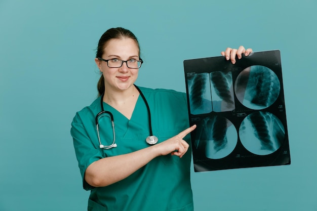 Giovane donna infermiera in uniforme medica con uno stetoscopio intorno al collo che tiene i raggi x del polmone che puntano con il dito indice su di esso guardando sorridente fiducioso in piedi su sfondo blu