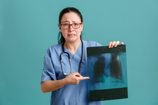 Giovane donna infermiera in uniforme medica con uno stetoscopio intorno al collo che tiene i raggi x del polmone che puntano con il dito indice verso di esso preoccupato e spaventato in piedi su sfondo blu