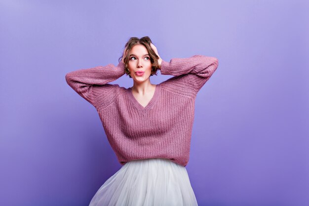 Giovane donna interessata in maglione oversize che tocca la sua testa sul muro viola. sognante ragazza dai capelli corti in gonna bianca che gode del servizio fotografico.